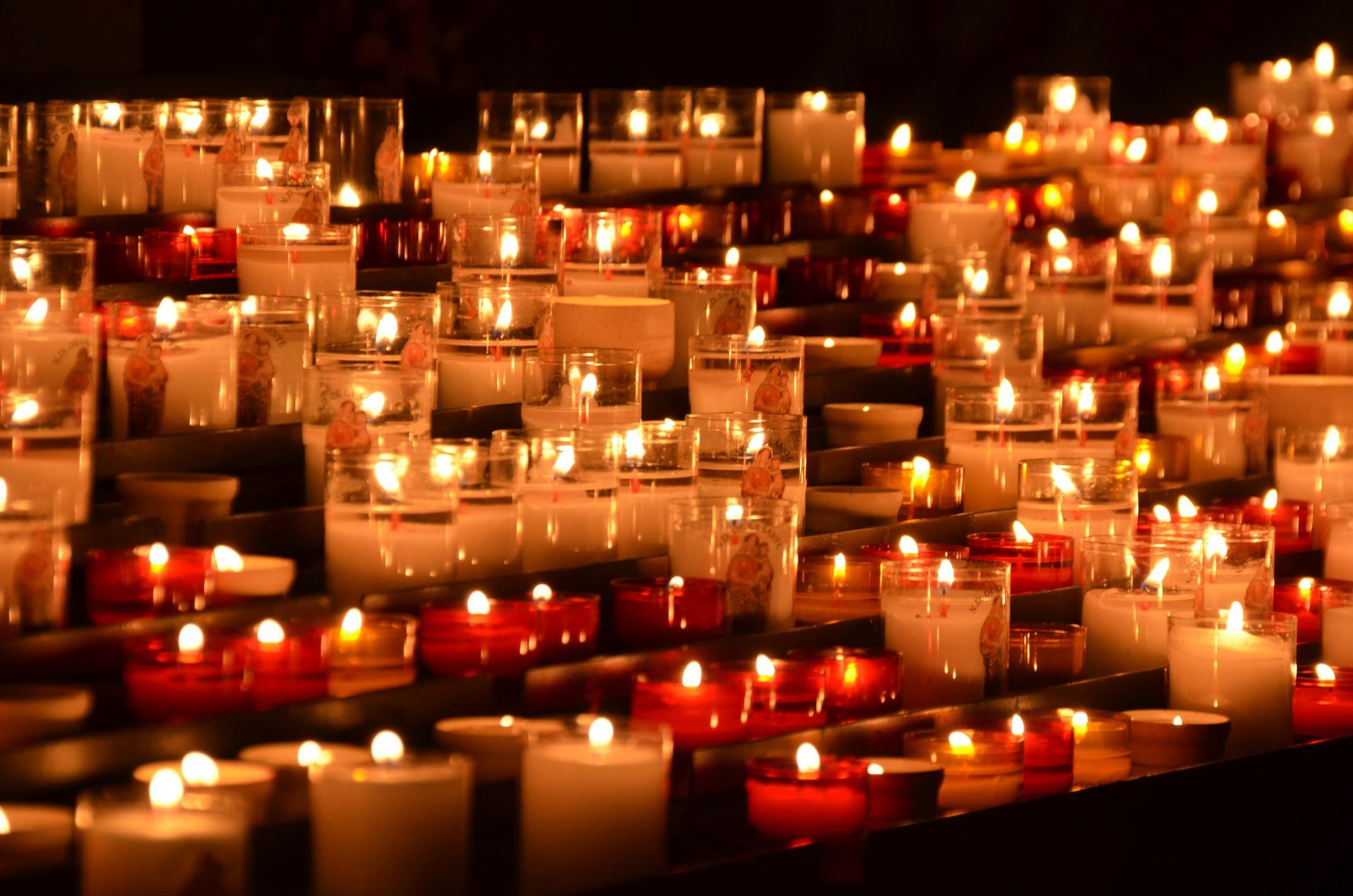 Rows of lit candles in glass holders, in shades of white and red.