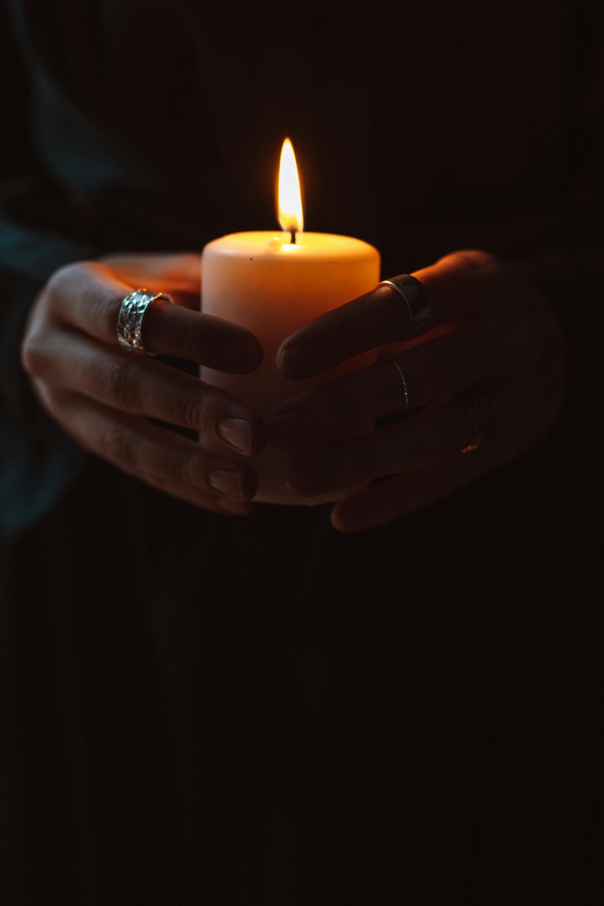 Hands holding a lit candle in a dark setting, rings visible.