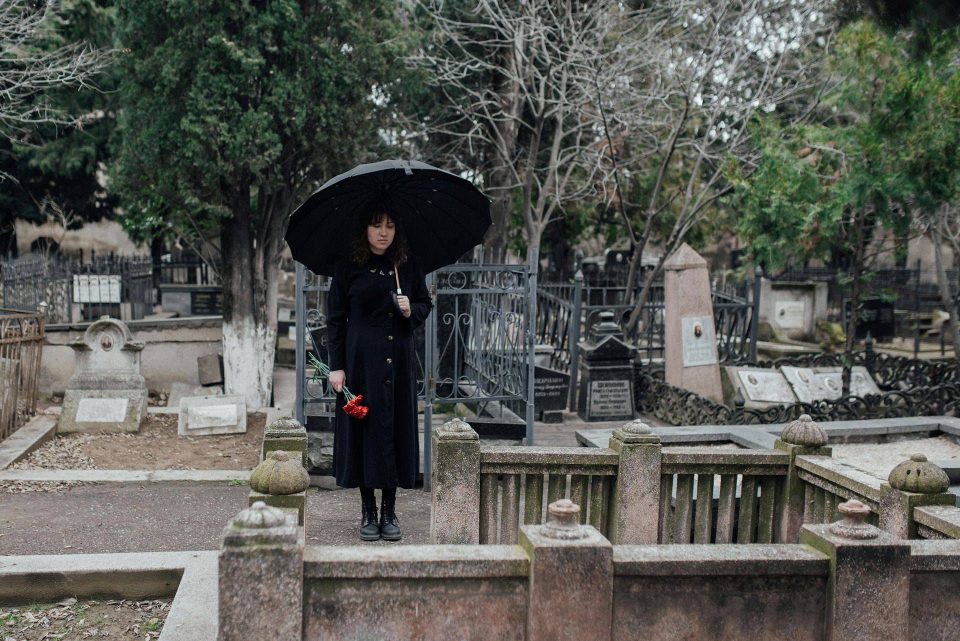 Woman in black coat and umbrella in a cemetery, holding a red flower.