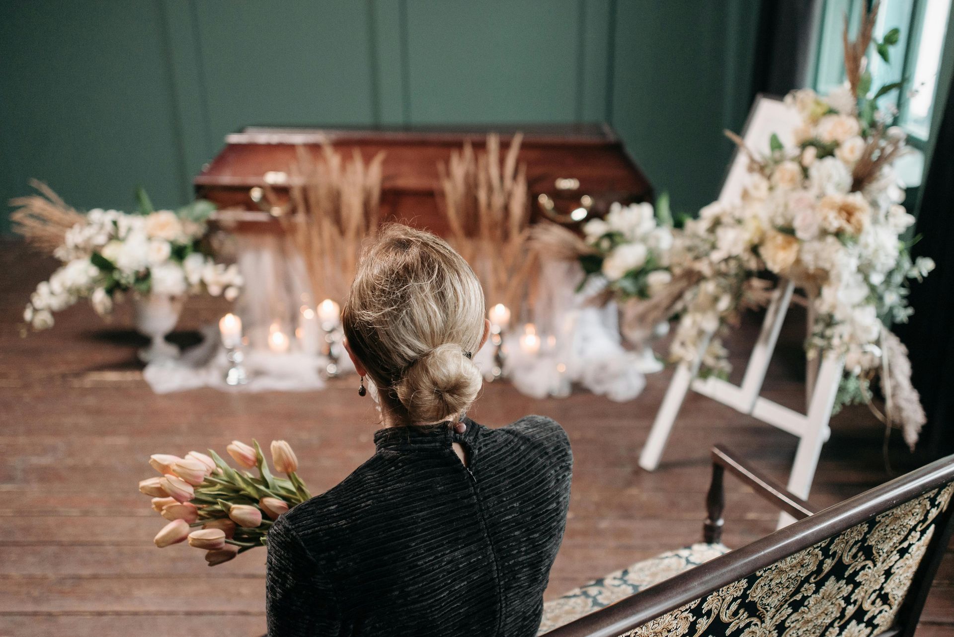 Woman holding flowers at a funeral, facing a coffin and floral arrangements.