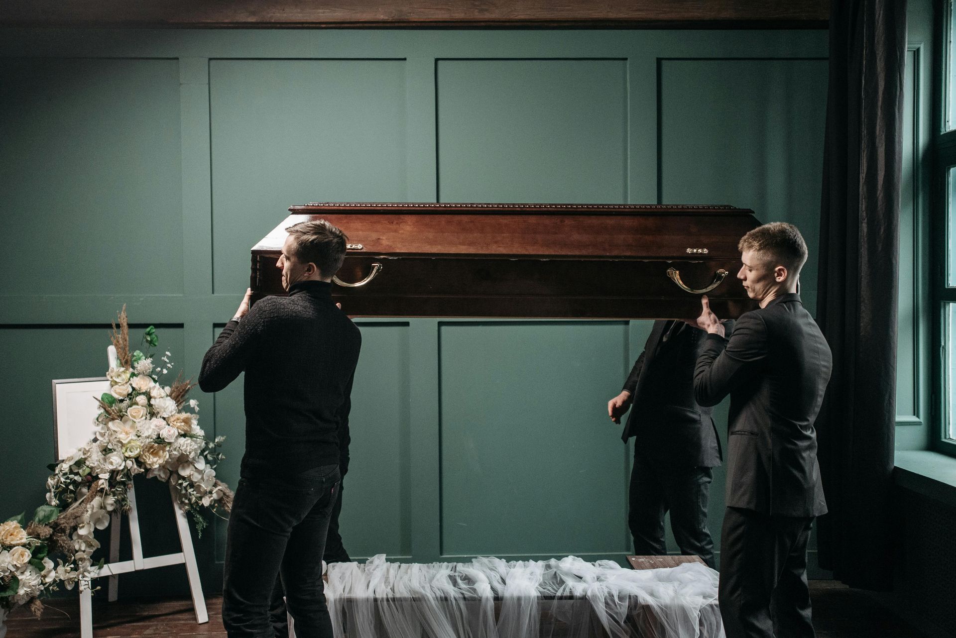 Pallbearers carrying a casket into a funeral home, near a floral arrangement.