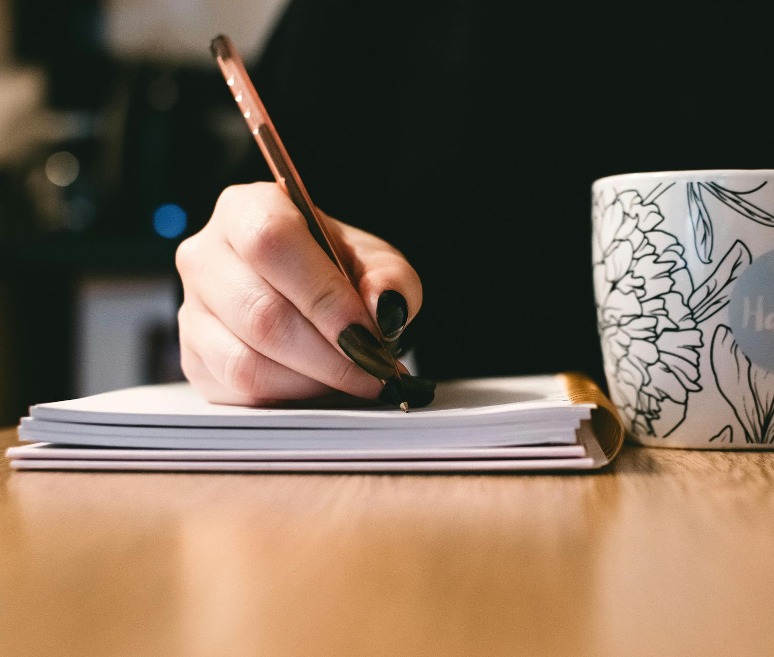 Hand writing in a notebook on a wooden table, next to a floral mug.