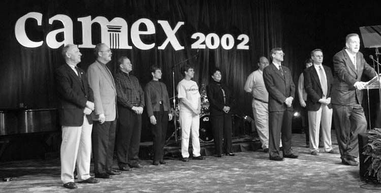 A group of men standing in front of a camex 2002 sign