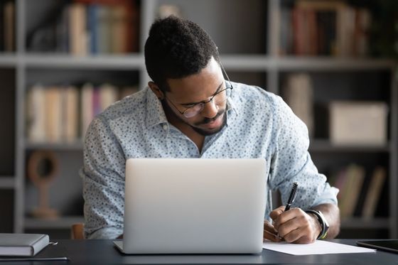 A man is sitting at a desk in front of a laptop computer writing on a piece of paper.