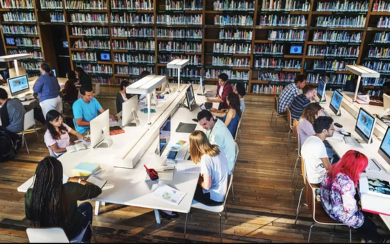 A group of people are sitting at tables in a library using computers.