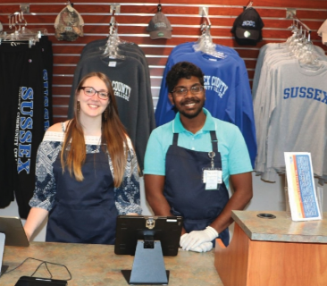 A man and a woman are posing for a picture in front of a wall of sussex shirts