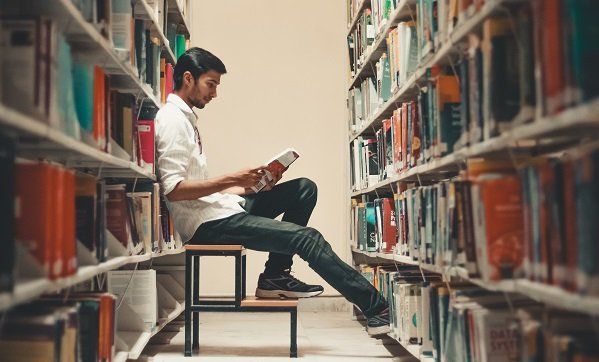 A man is sitting on a stool in a library reading a book.