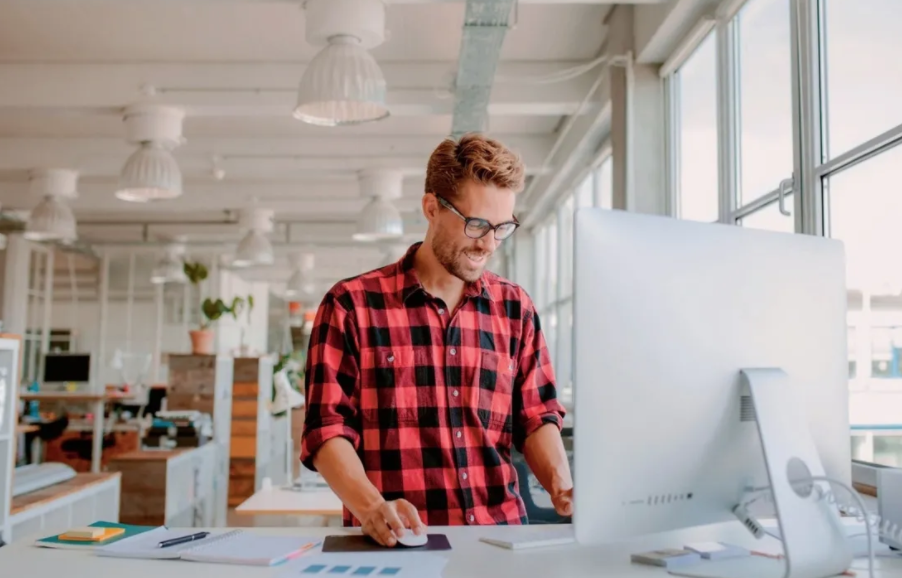 A man in a plaid shirt is standing in front of a computer in an office.