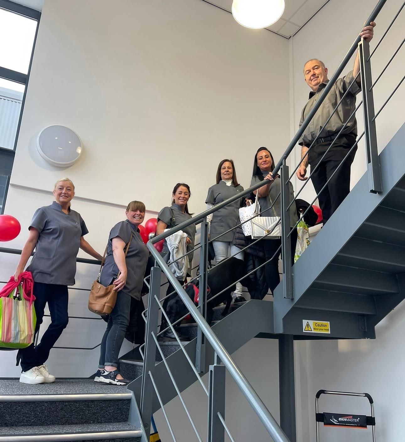 Six people on a staircase. Most wear grey uniforms. One man at the top smiles, holding the railing.