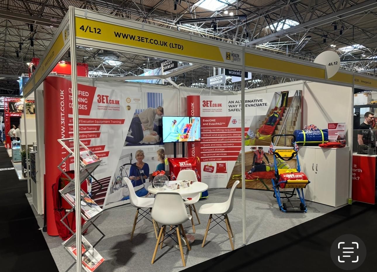 An exhibition booth displays products and brochures. Chairs and a table are set up for visitors.