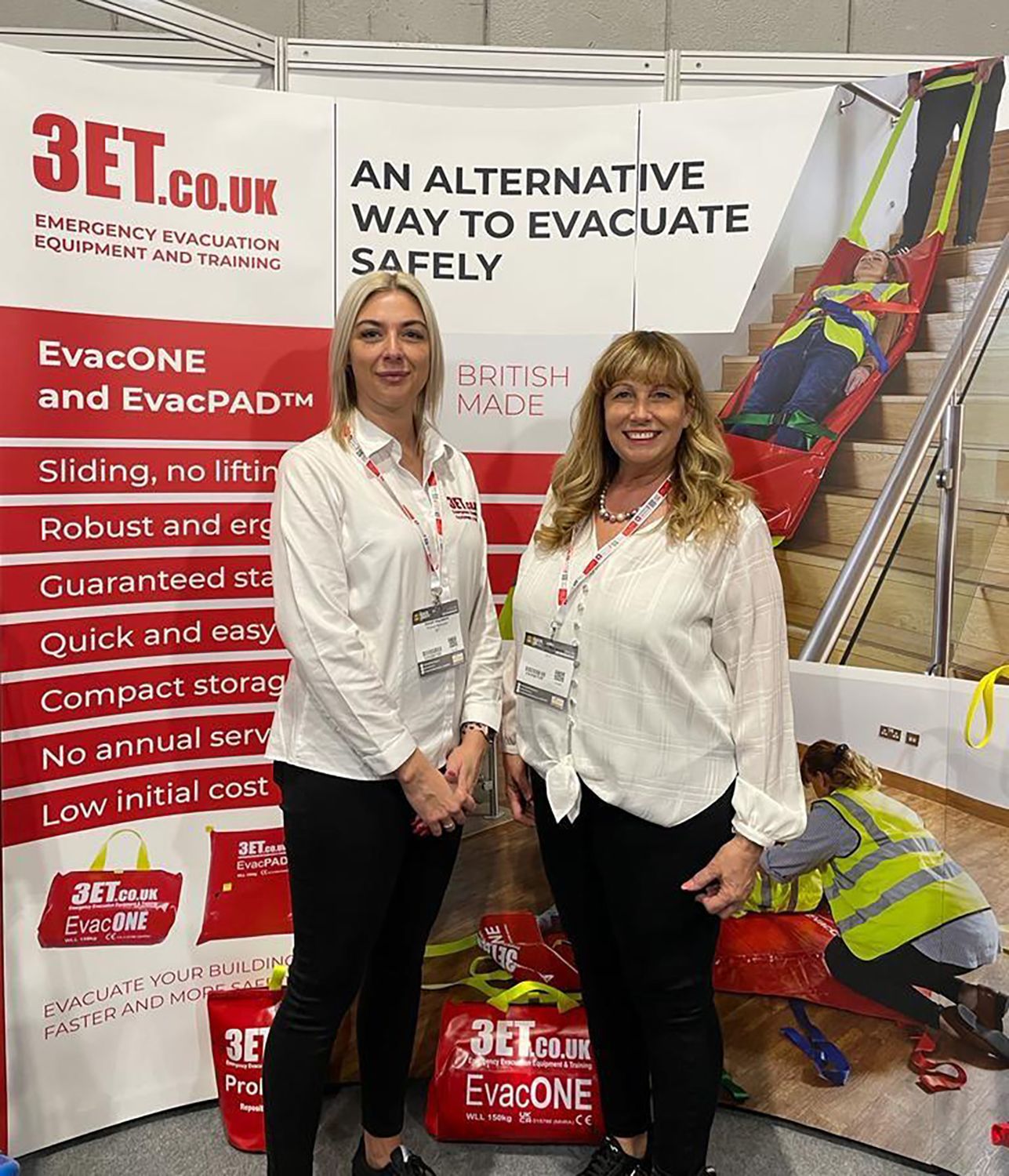 Two women stand at a booth promoting a stair evacuation system. Signage displays the product name, features, and website.