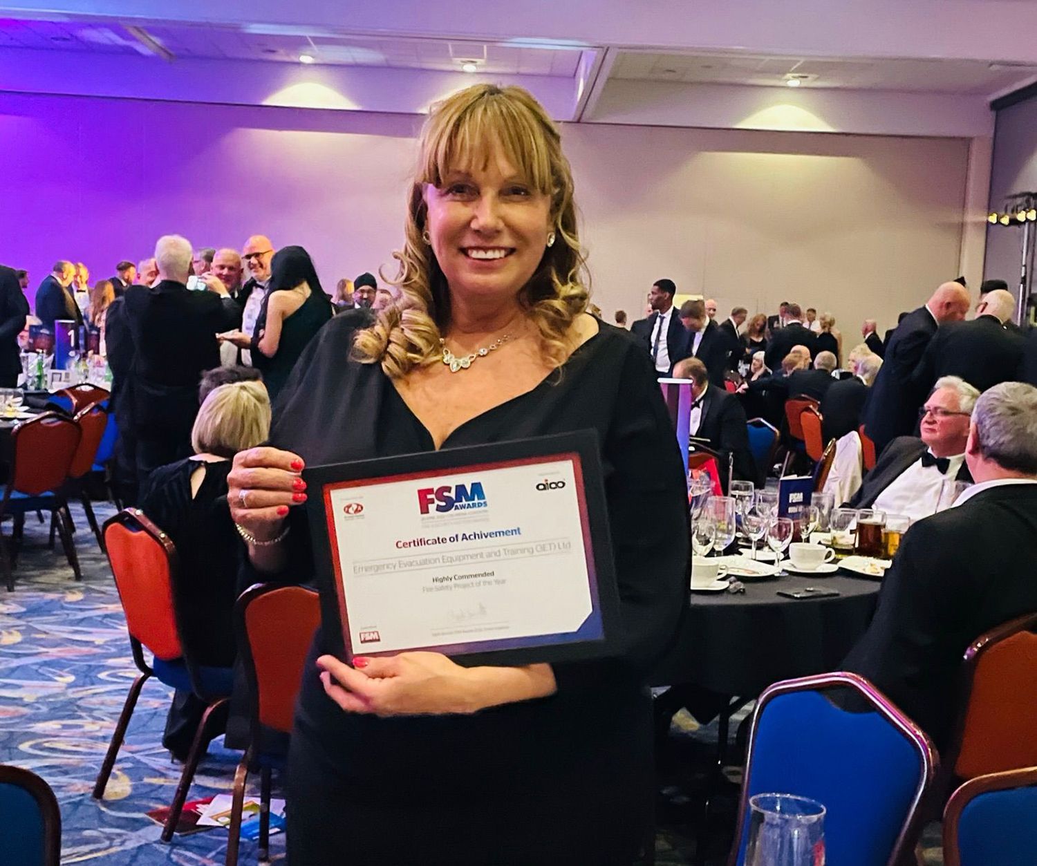 Woman in black dress smiling, holding award at formal event. Large banquet hall in background.