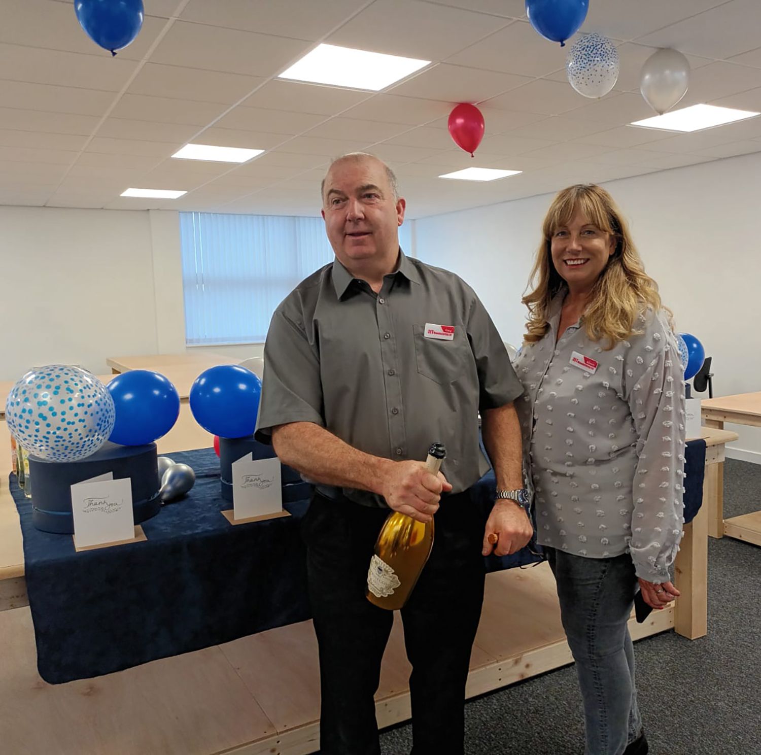 Man and woman at event with balloons, man holding champagne, and a table with items.
