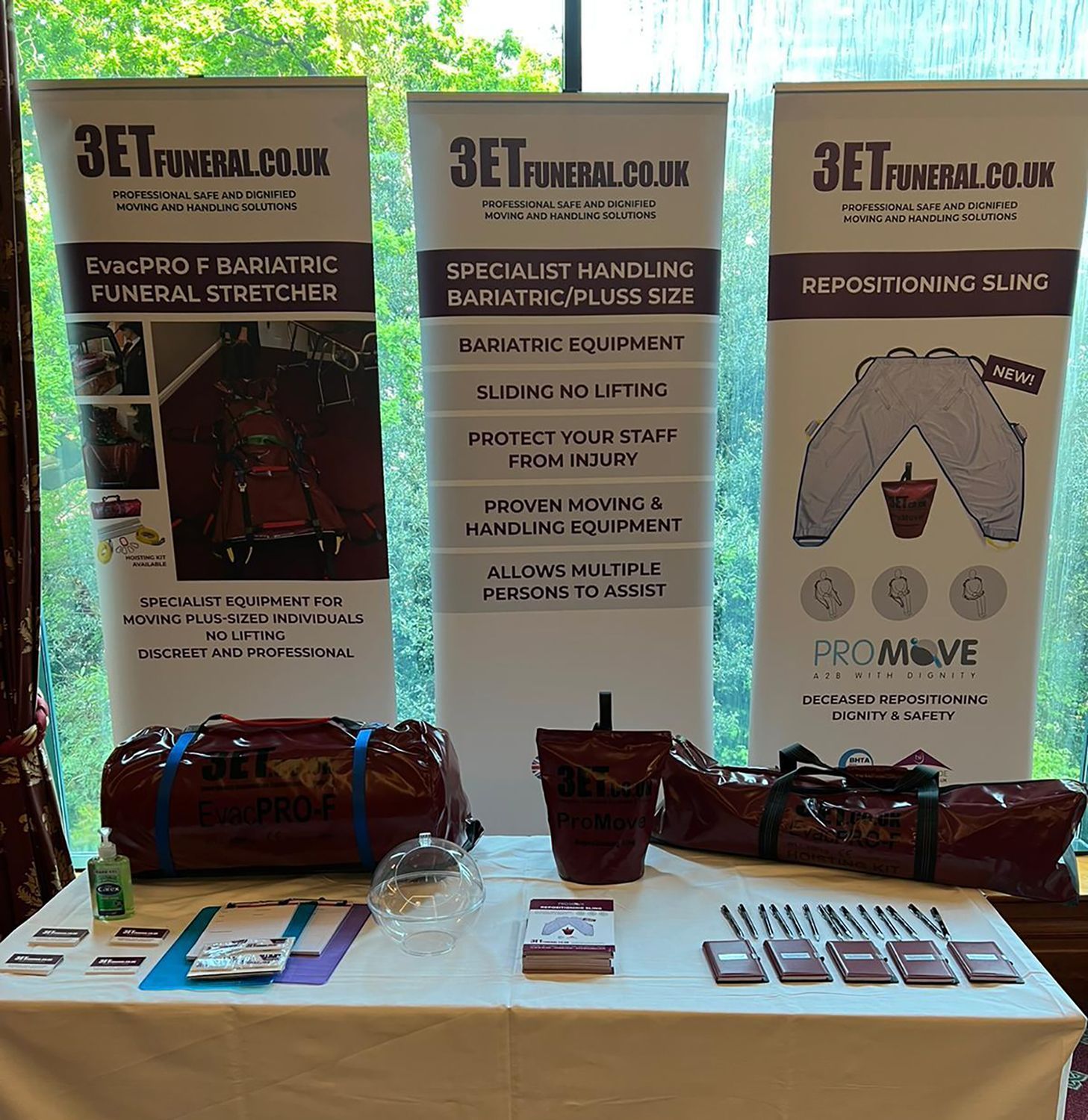 Funeral equipment display: banners, stretcher, repositioning aid, bags, promotional materials, white table.