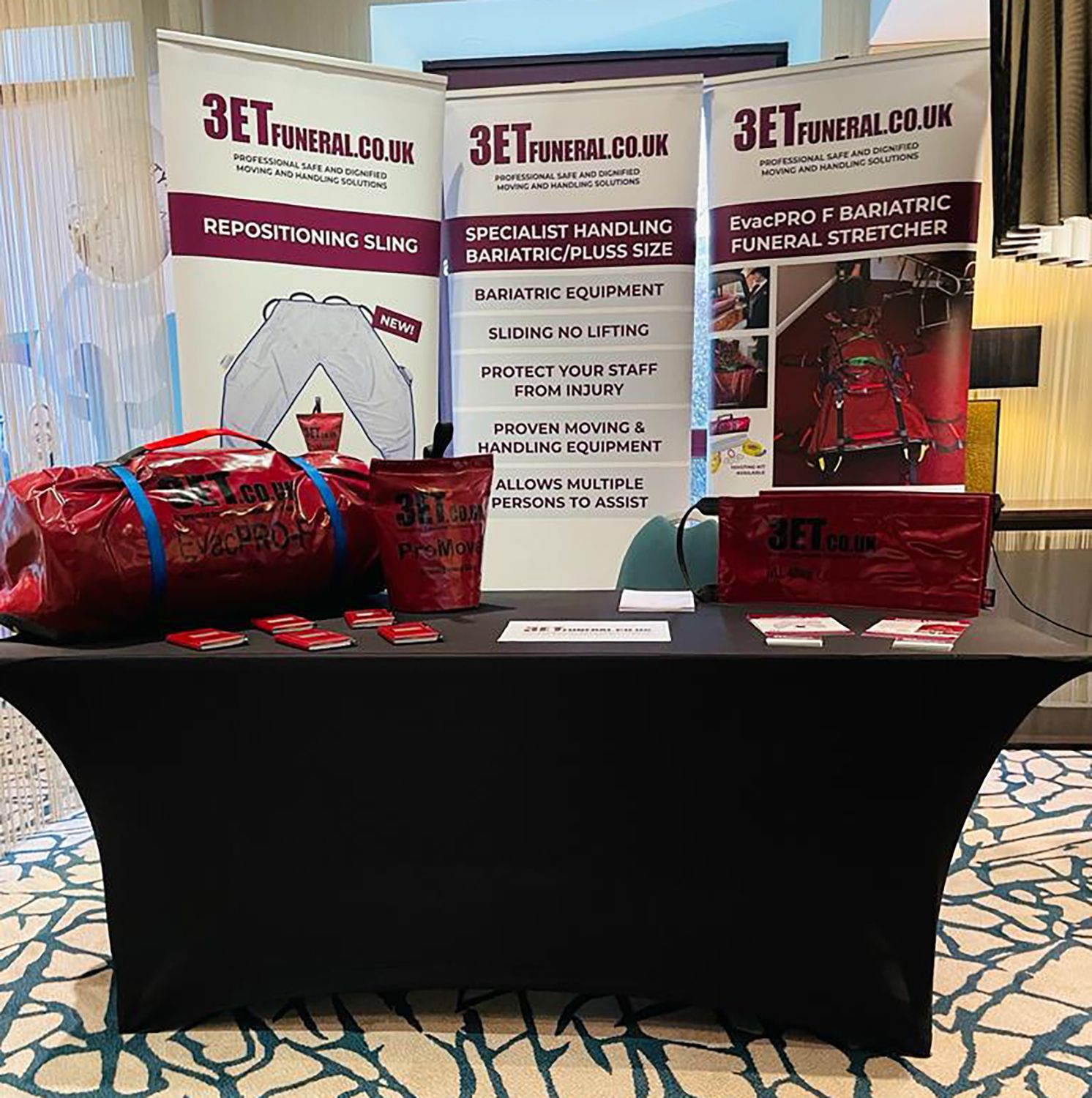 Trade show display: red bags, brochures, and banner stands for 3ET FUNERAL.CO.UK on a black table.