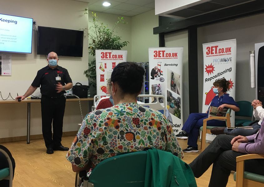 A man in a mask presents to a seated group, likely in a hospital setting. The man is pointing.