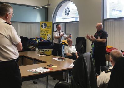 Men in uniform at a table, discussing equipment. One holds object, gesturing. Meeting in a room.