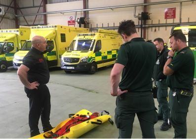 Emergency workers in green uniforms examine a stretcher in an ambulance bay, ambulances in background.