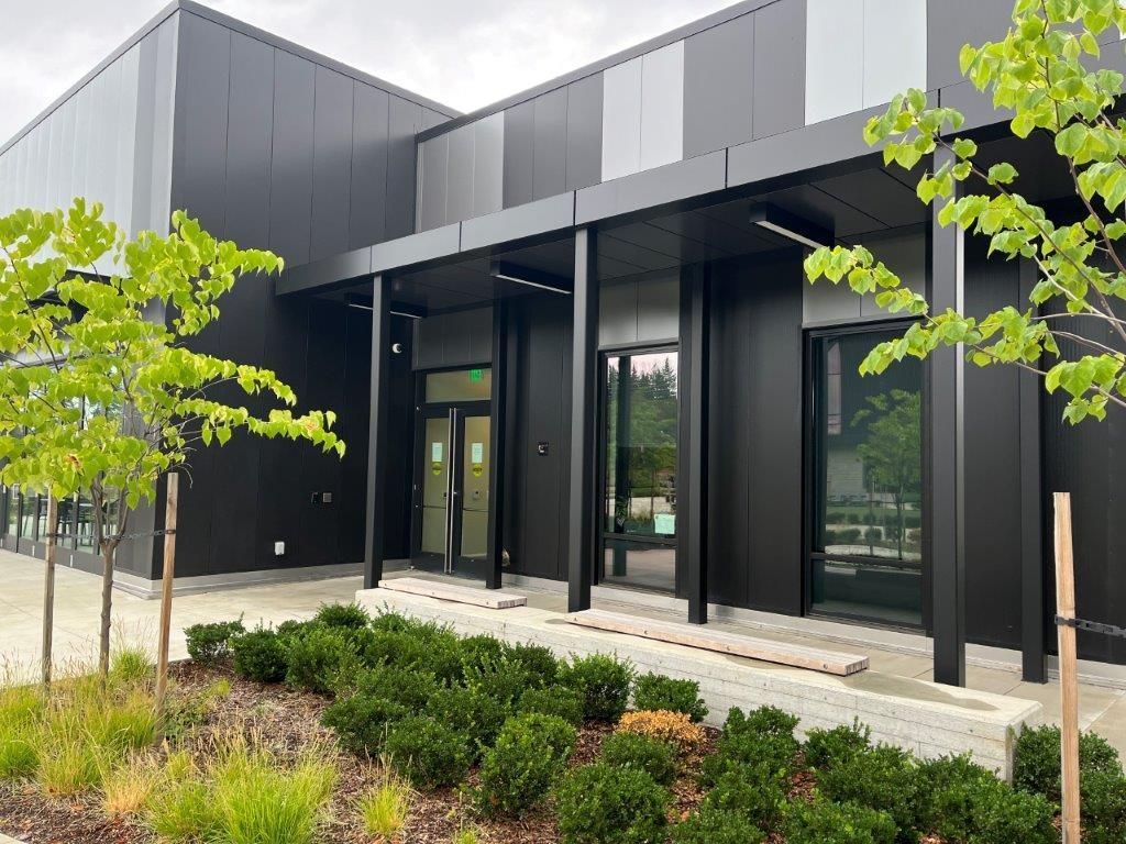 Modern black building with large windows, trees, and landscaped shrubs along the entrance