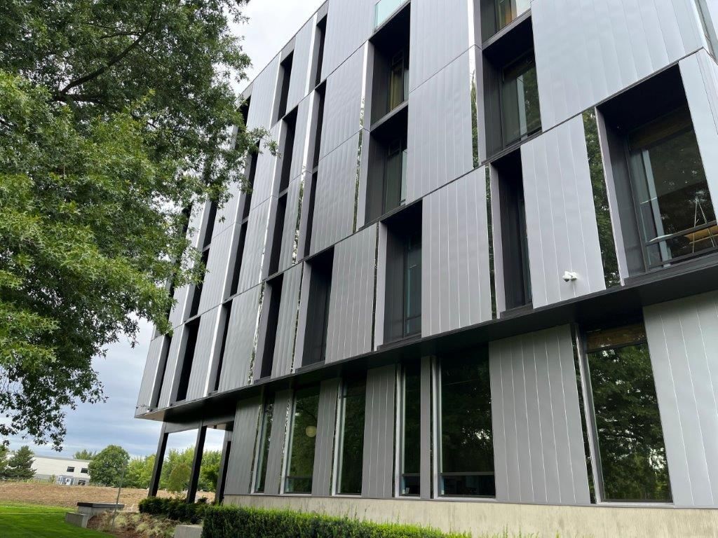 Modern building with dark vertical panels and tall windows beside leafy trees and open field