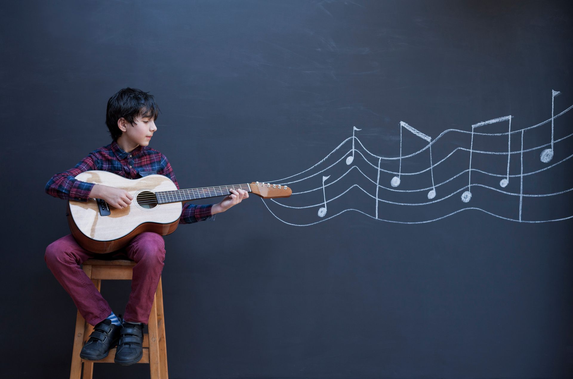 A young boy is sitting on a stool playing an acoustic guitar.