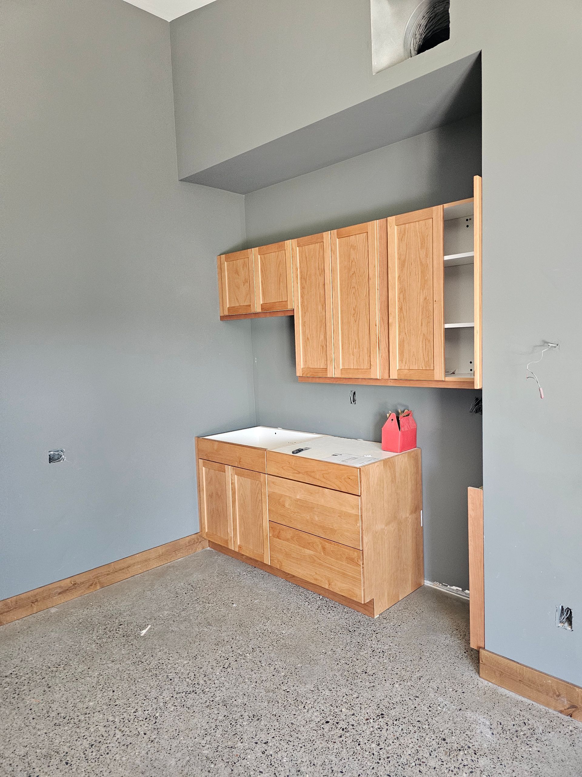 Cabinets in a room under construction with gray walls, unfinished floor, and a red toolbox.