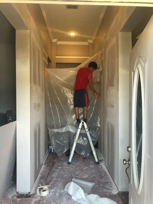 Person on ladder working on drywall in a hallway, plastic sheeting covering the work area.