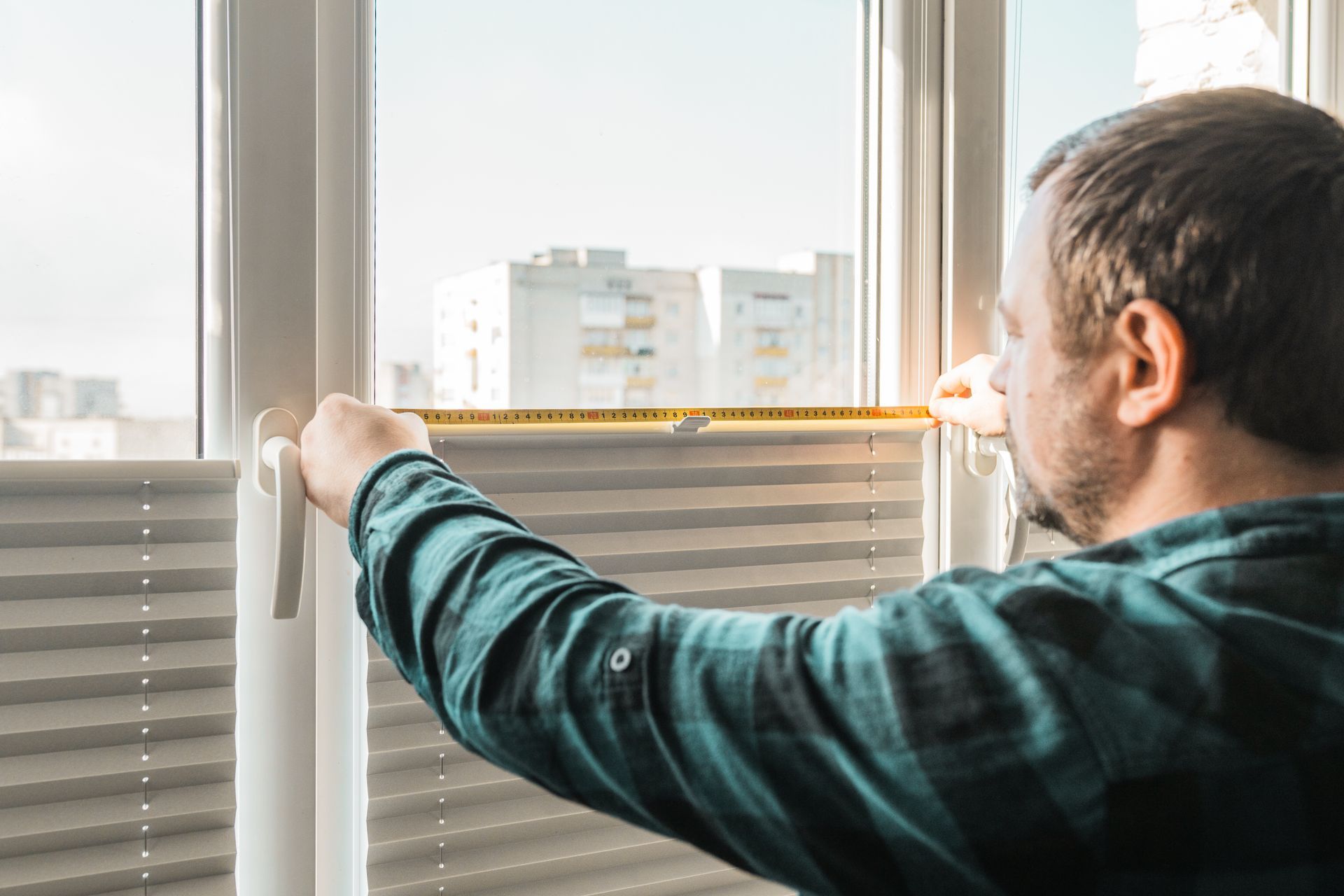 Man measuring a window for blinds with a yellow tape measure, sunny interior setting.