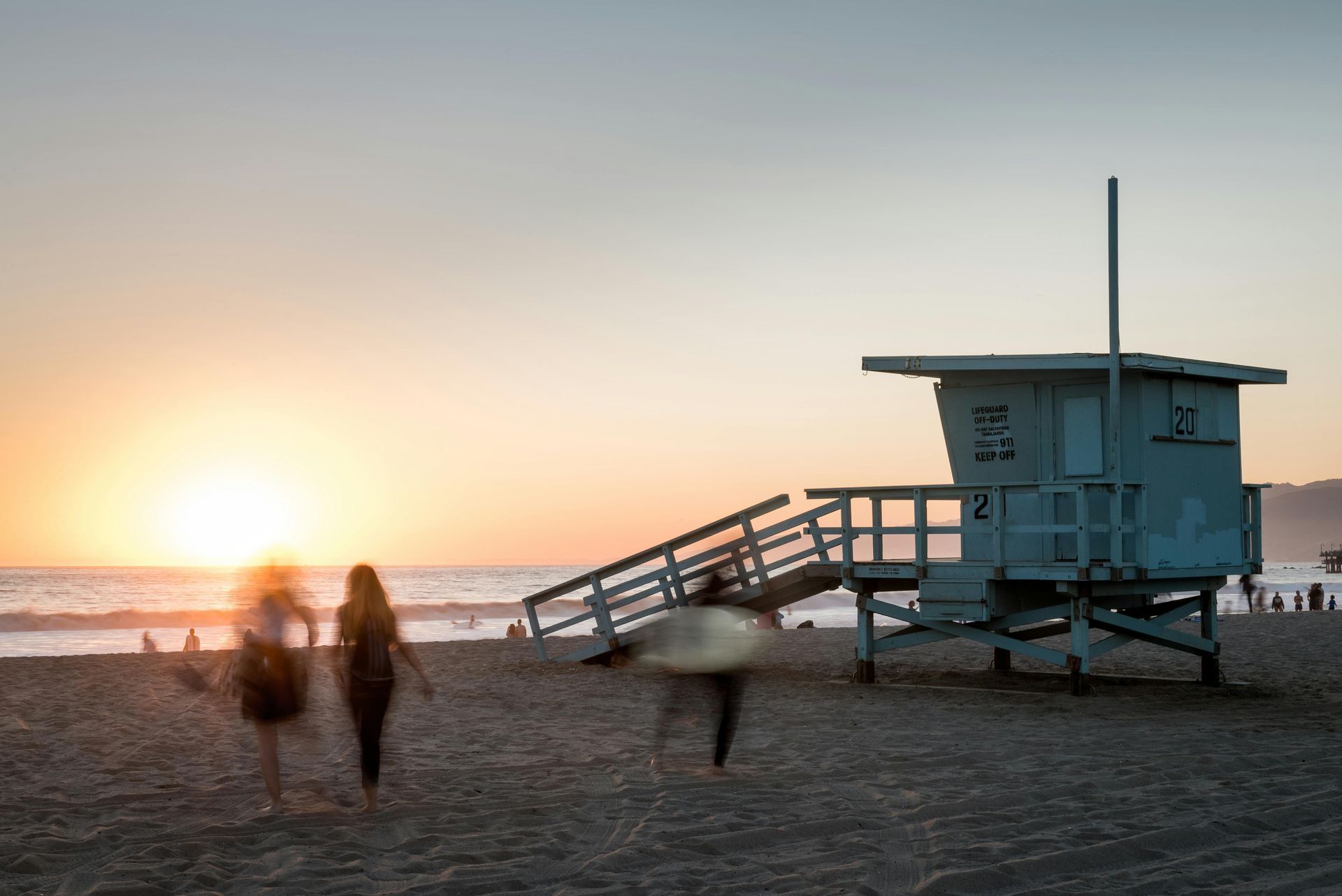 A group of people are walking on a beach near a lifeguard tower at sunset.