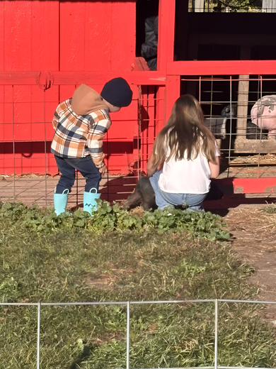 Two children near a red pen. One in blue boots and a plaid shirt crouches. The other sits, facing the pen.