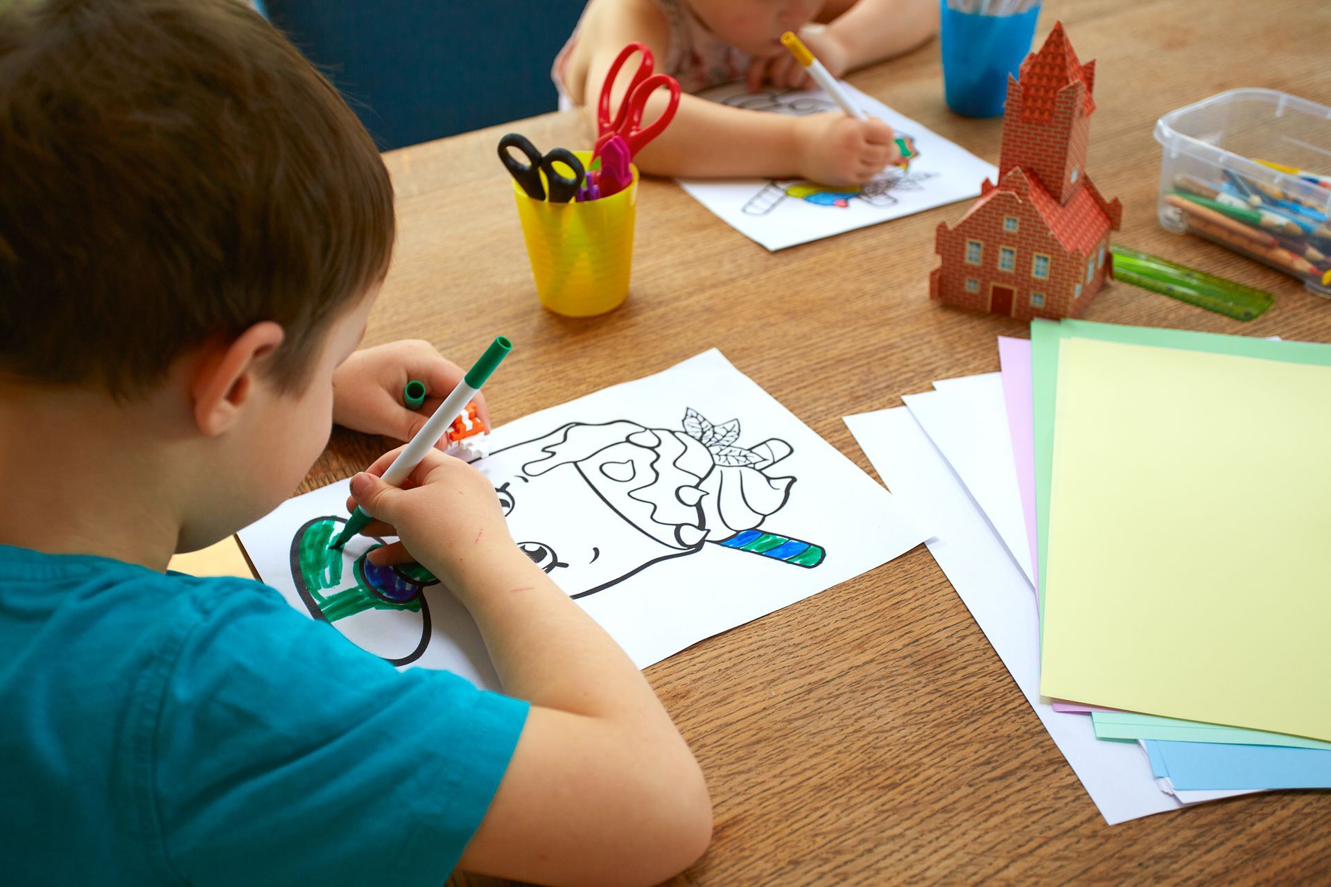 Children coloring at a table with crayons, scissors, and a paper craft house.