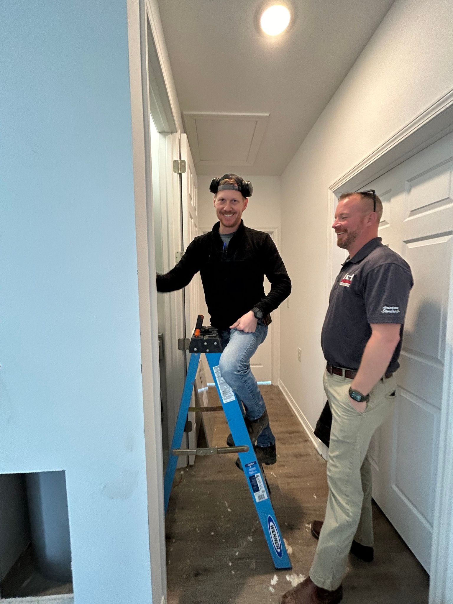 Two men are standing next to each other in a hallway . one of the men is sitting on a ladder.