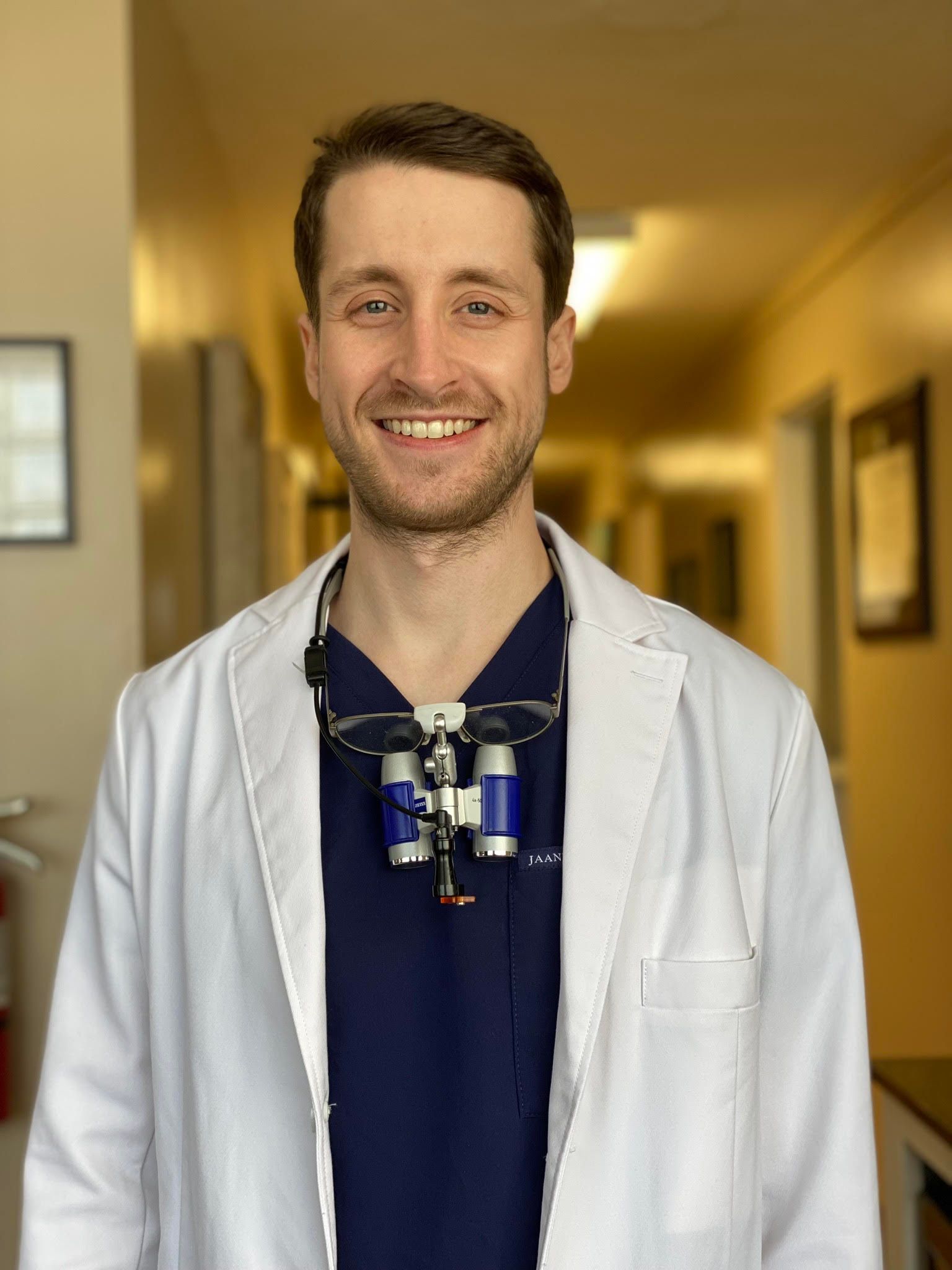 A dentist wearing a white coat and a blue scrub is smiling in a hallway.