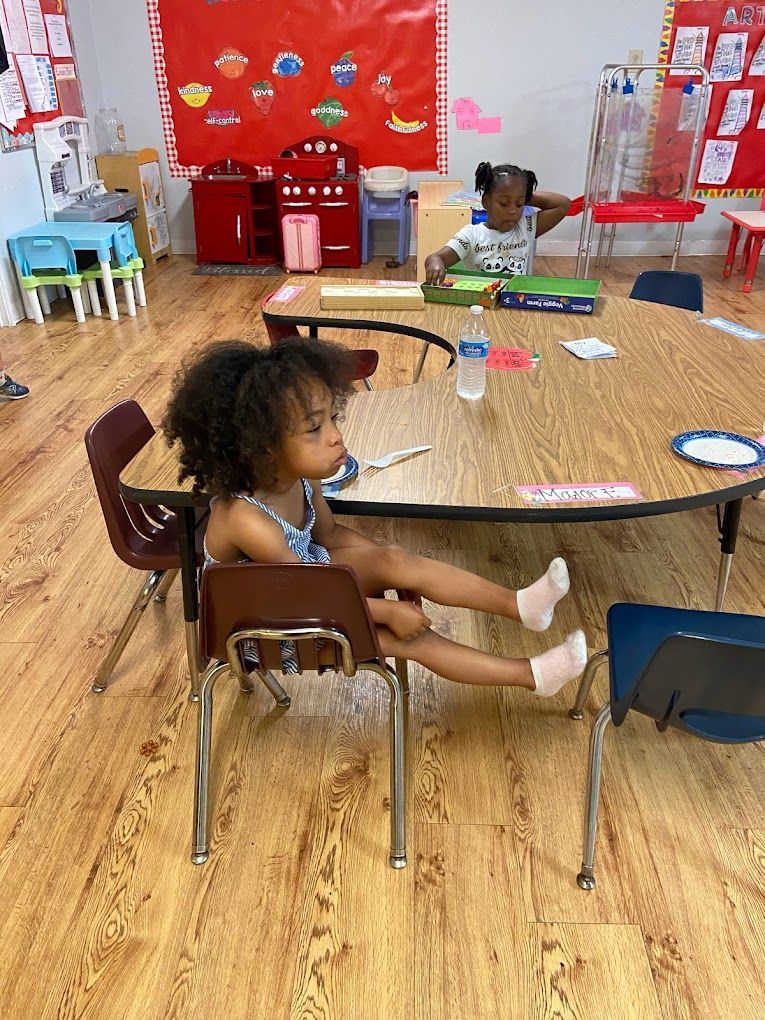 A little girl is sitting at a table in a classroom with her legs crossed.