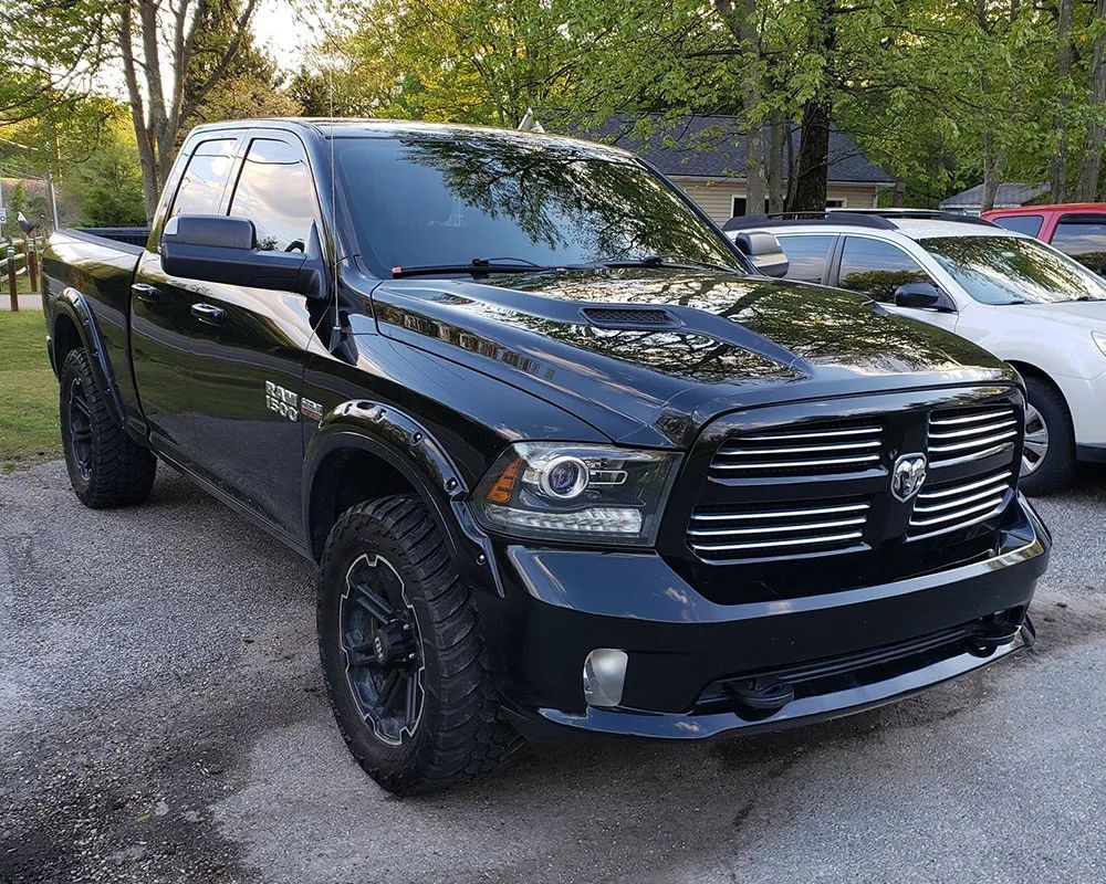 Black Ram 1500 truck parked on a paved surface. The truck is in front of a building with a white car in the background.