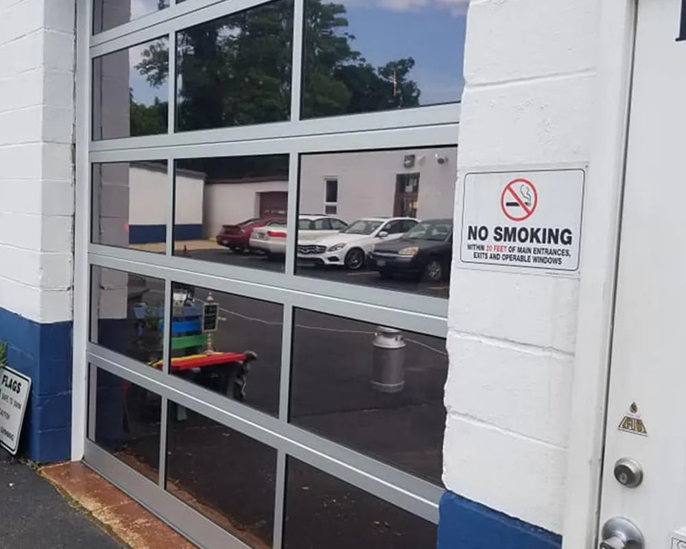 Glass garage door with a No Smoking sign, reflecting parked cars and a tree-lined area.