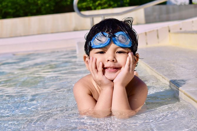 Photo of boy wearing blue goggles laying prone in shallow swimming pool resting his face in his hands and smiling at the camera.