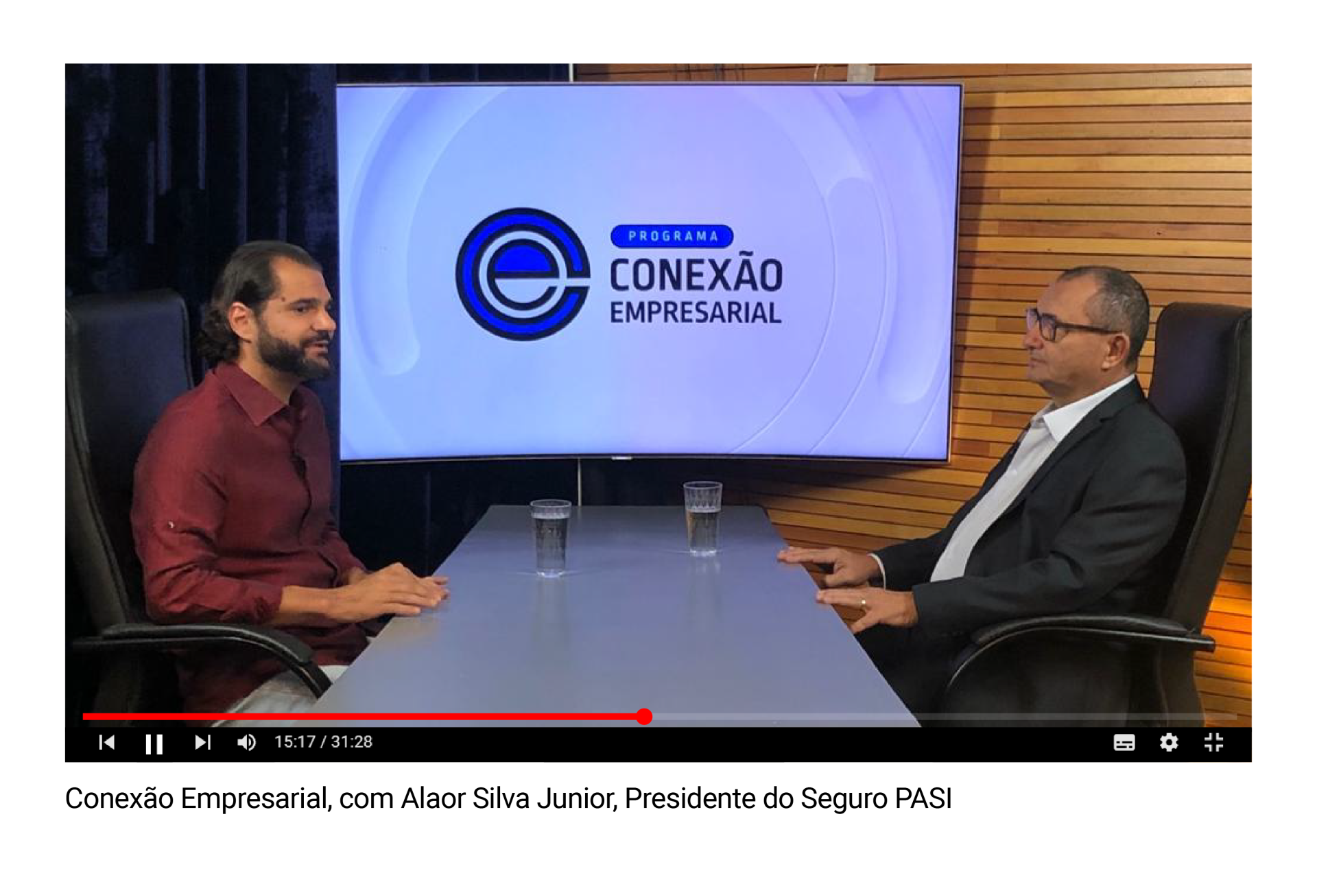 Dois homens estão sentados à mesa conversando entre si em frente a uma tela grande.