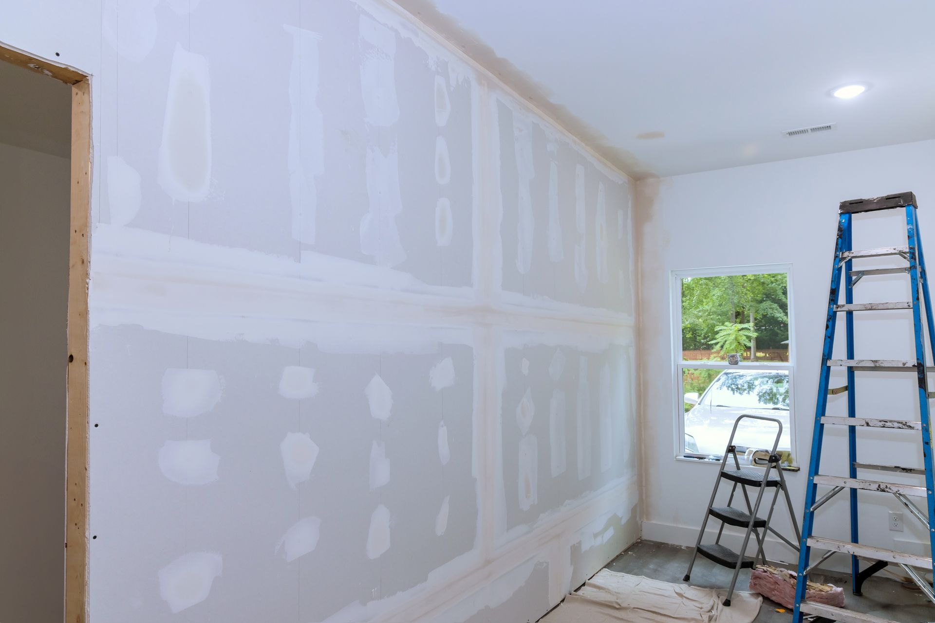 Freshly mudded drywall in a room, with ladders and tools near a window.