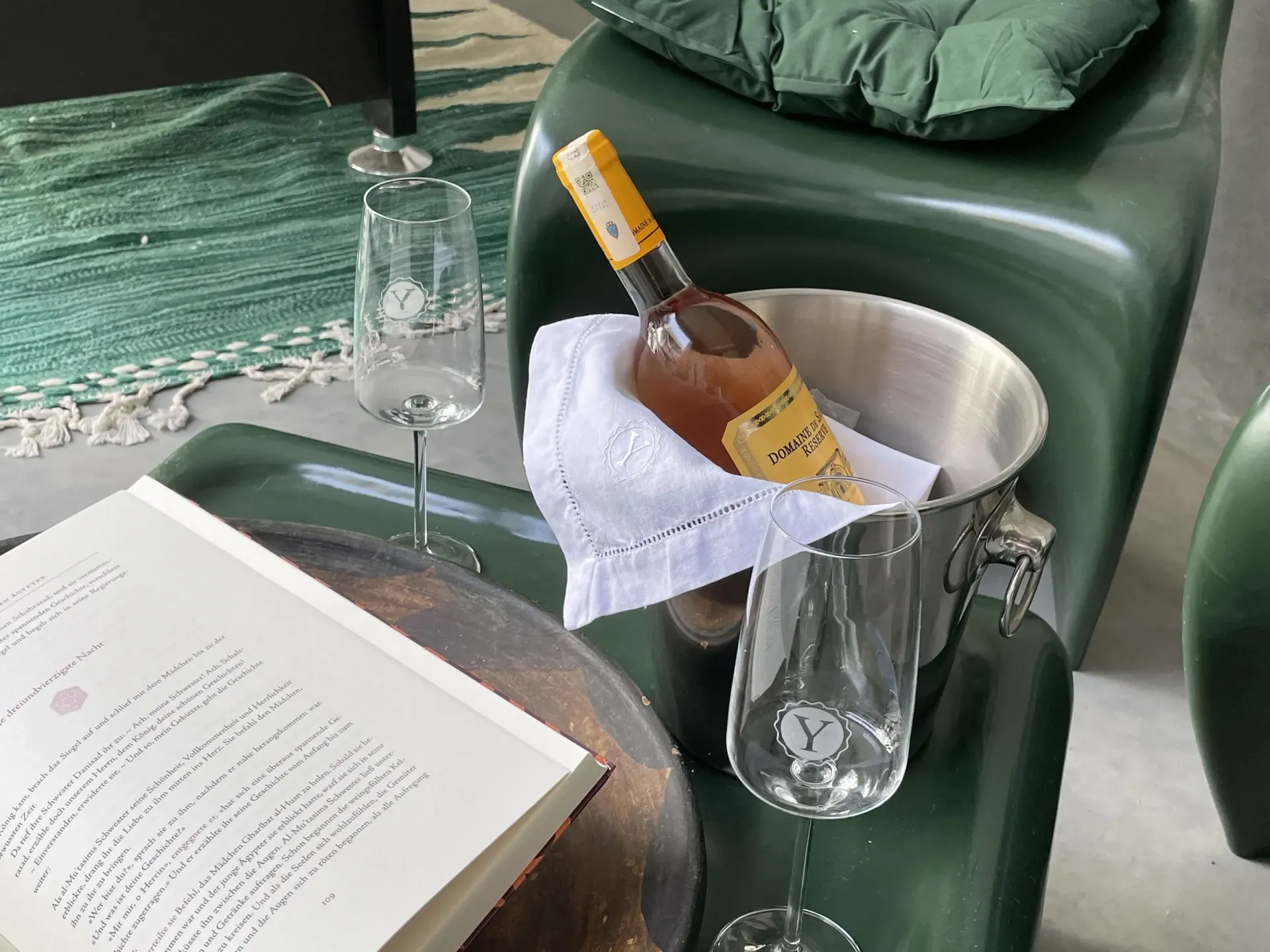 A bottle of wine is in a bucket on a table next to a book and wine glasses.