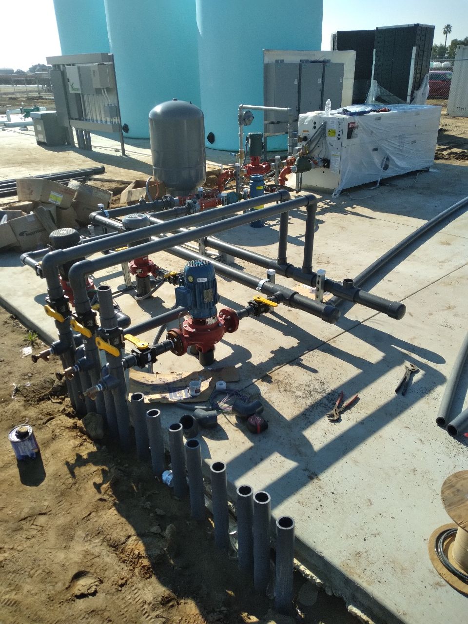 Piping and pumps at a construction site with blue tanks in the background.