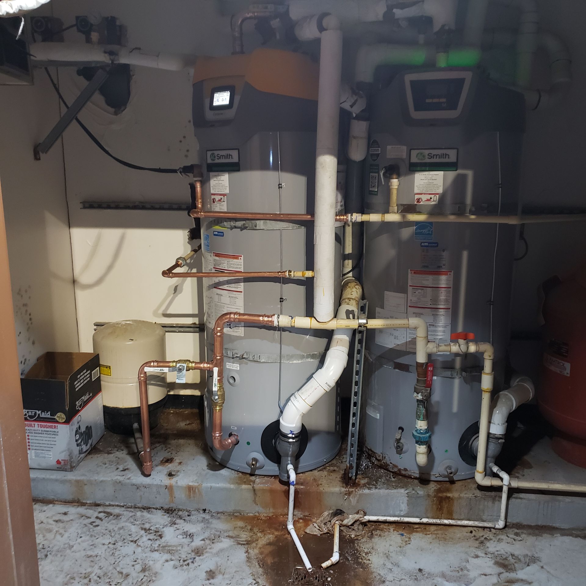 Two water heaters with copper and PVC piping in a utility room. The floor shows water stains.