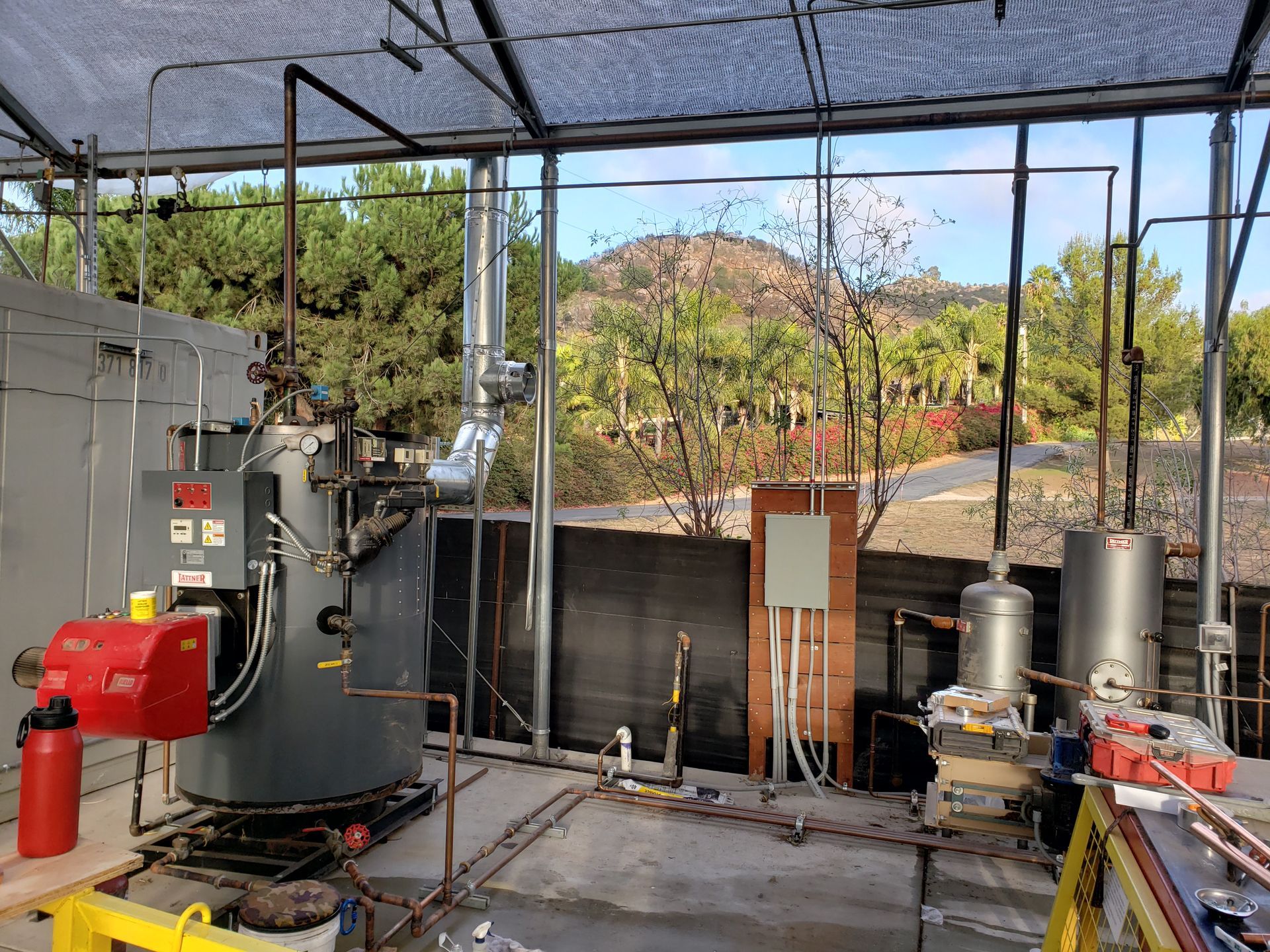An outdoor mechanical setup with a large boiler, two water heaters, and copper pipes.
