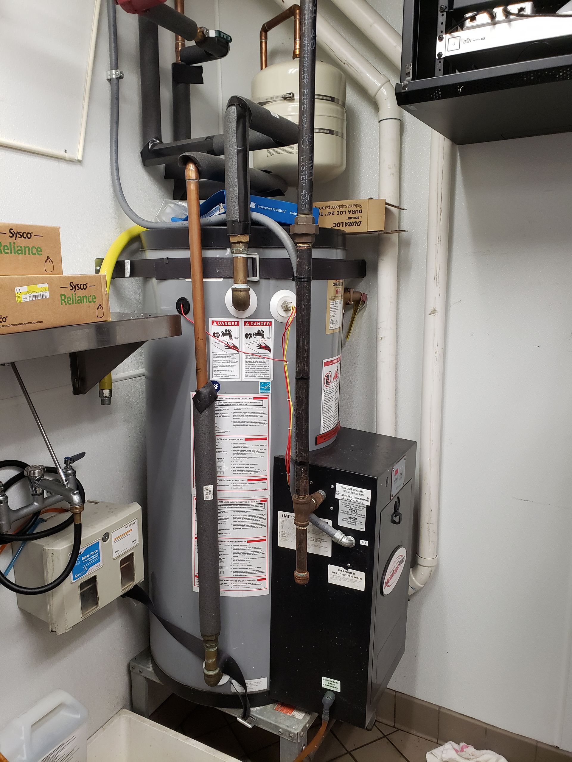 A water heater in a commercial kitchen. It has pipes, labels, and a black panel.