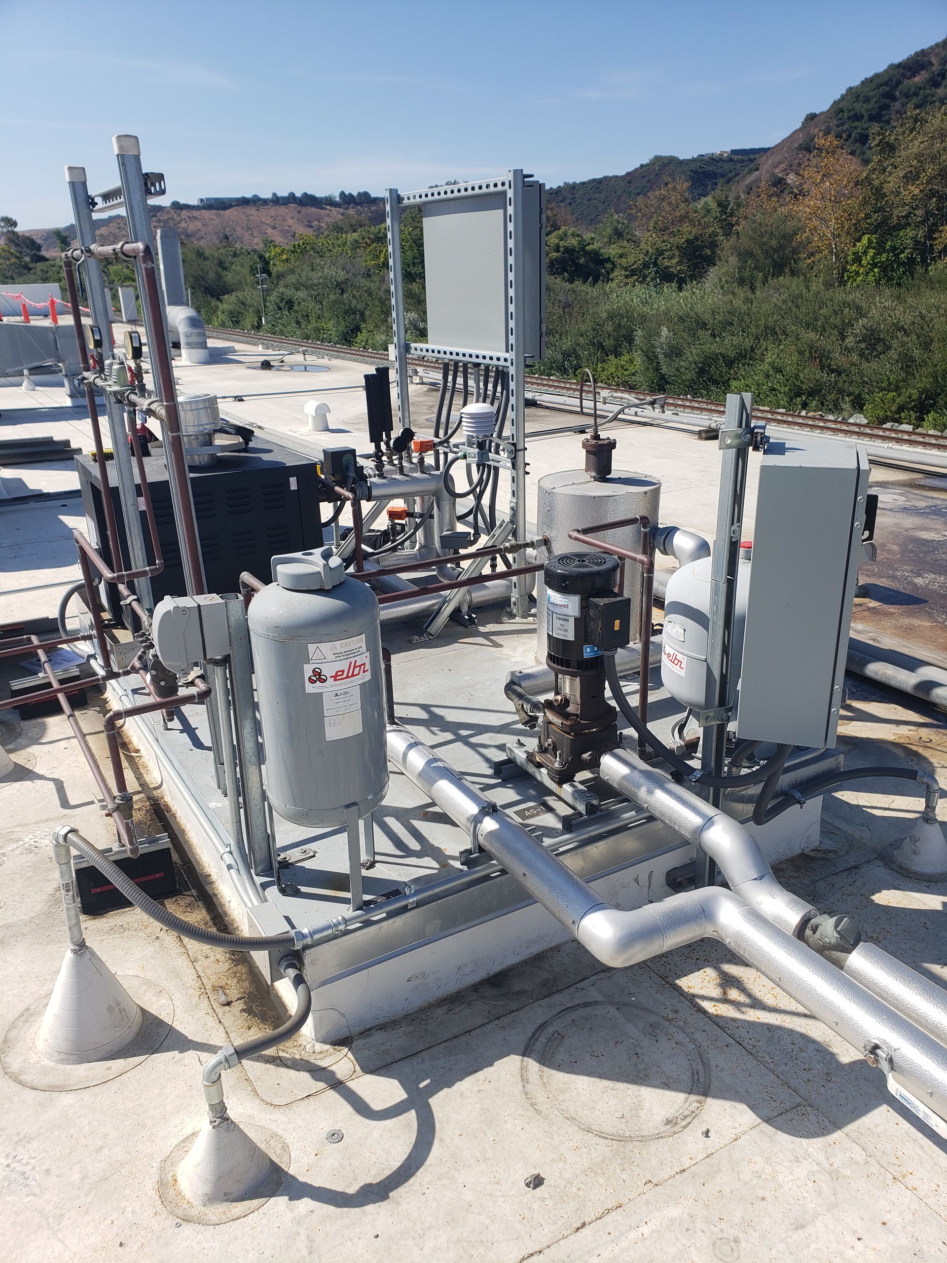 Industrial rooftop water pump and control system. Gray tanks and pipes are mounted on a platform.