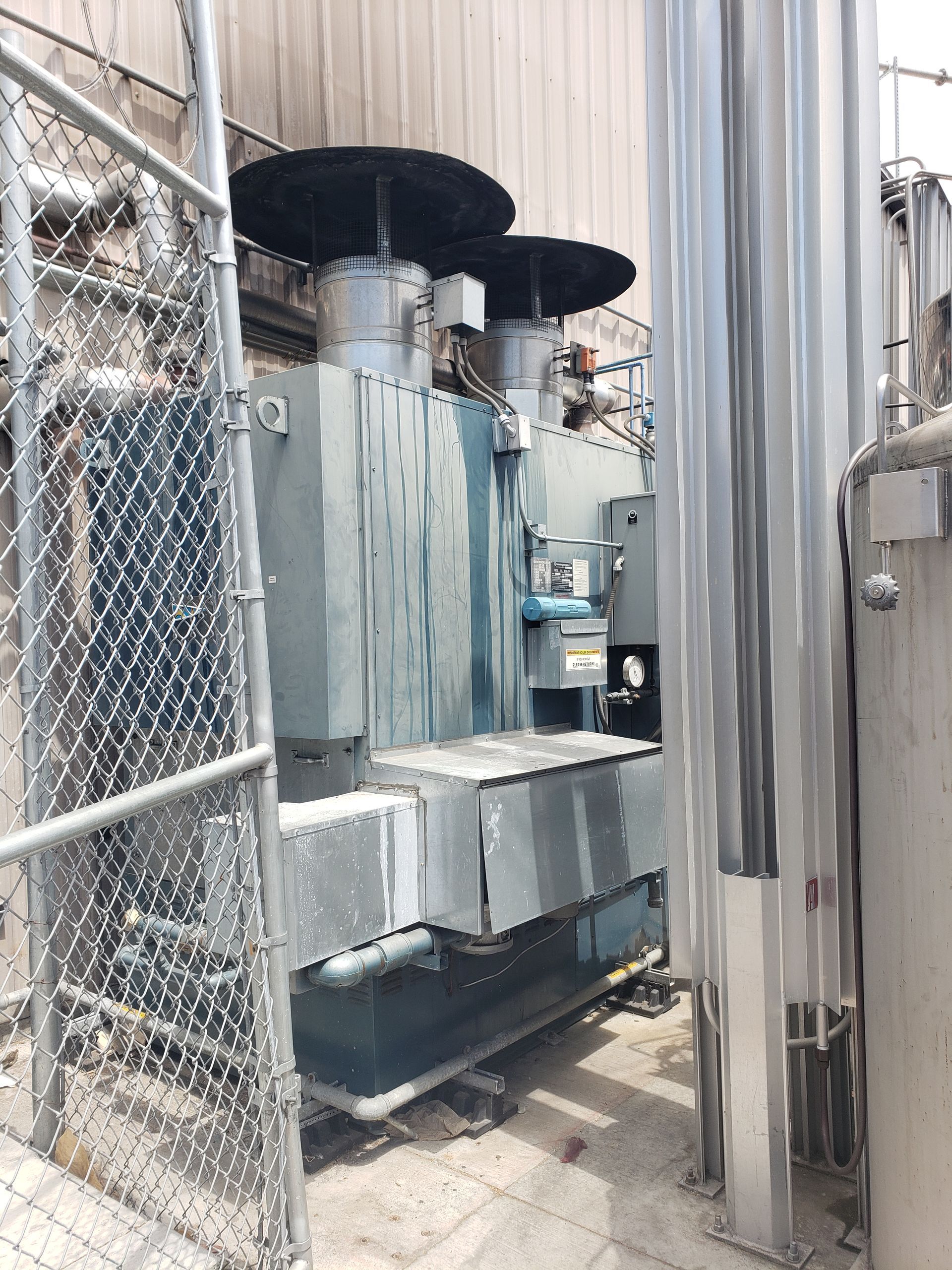 Large, gray electrical transformer outside a building, behind a chain-link fence.