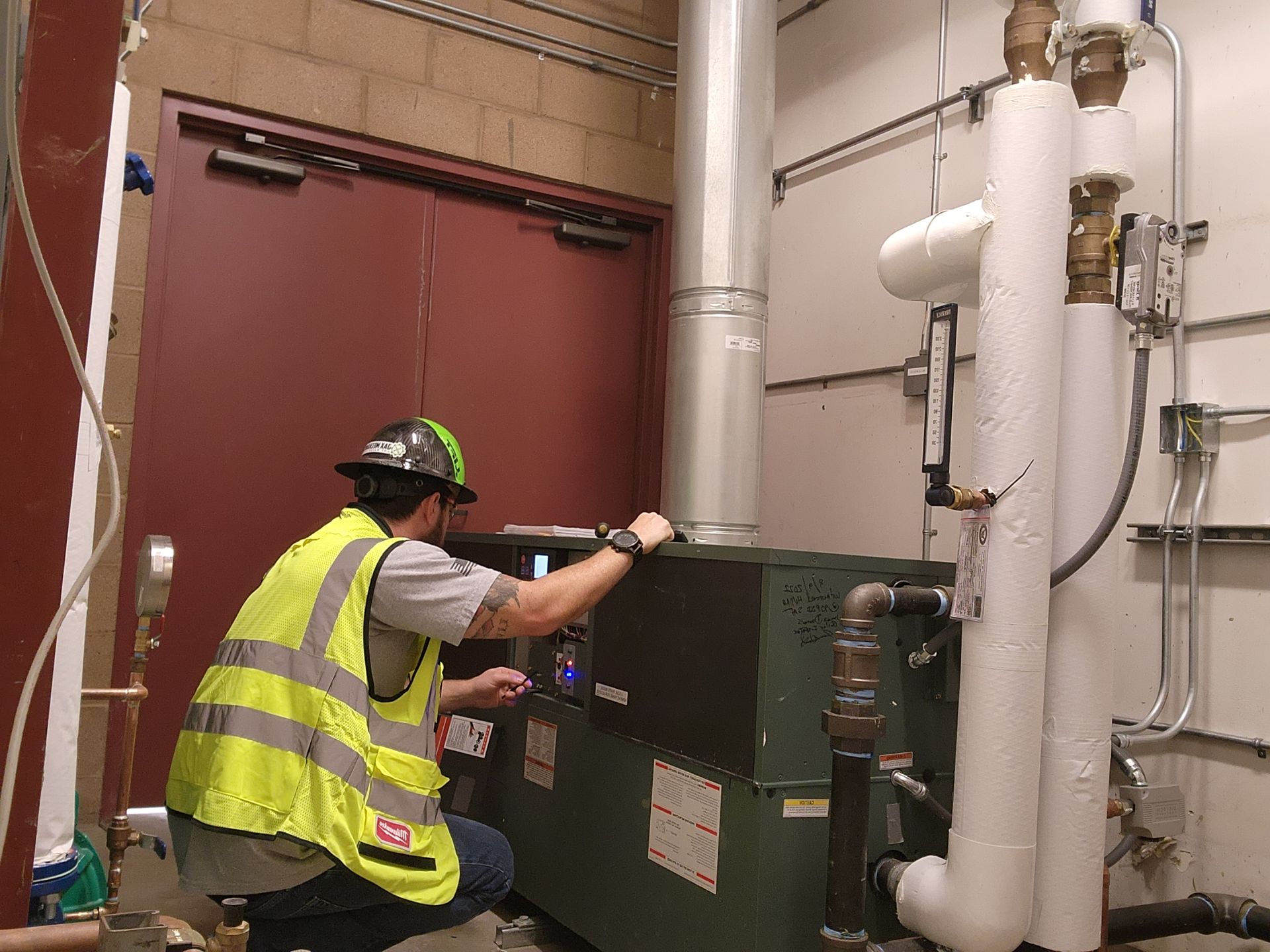 A person in a yellow safety vest and hard hat inspects a machine in a utility room.