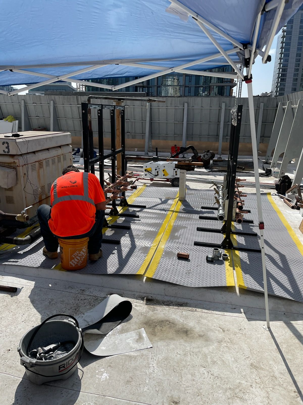 Construction workers on a rooftop assembling plumbing fixtures. One worker in an orange vest sits near a bucket.
