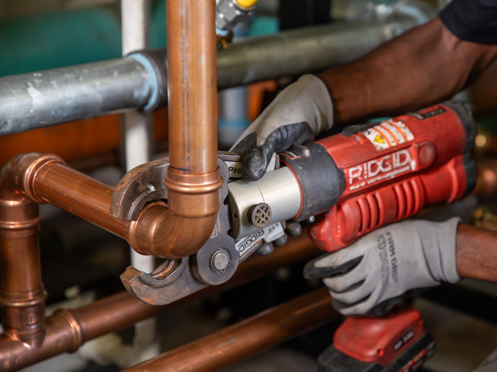 Plumber using a red Ridgid press tool to connect copper pipes, wearing gloves, in a mechanical room.