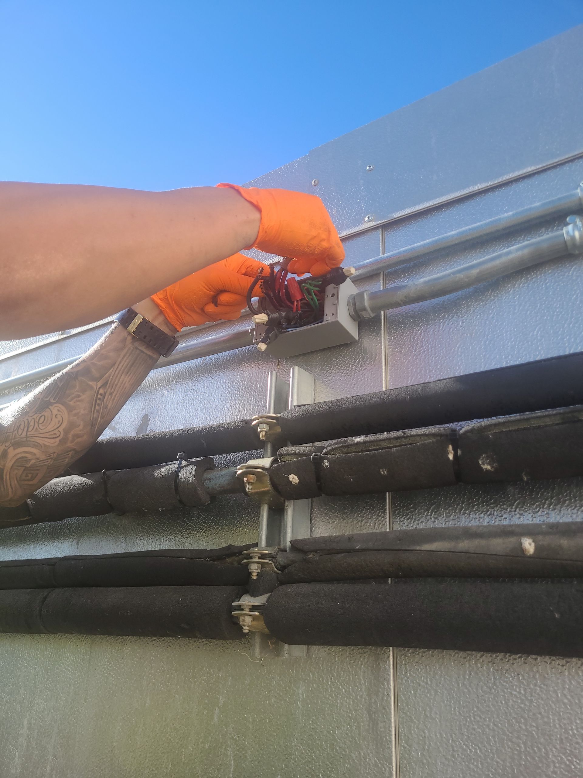 Person in orange gloves working on pipes outdoors against a metal wall and blue sky.